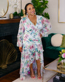A woman models a white high-low floral dress with pink, green, and blue watercolor-style flowers in a decorated living room setting.