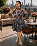 A woman models a knee-length floral dress with sheer long sleeves and a high neckline, standing on a rooftop patio with a city skyline in the background.